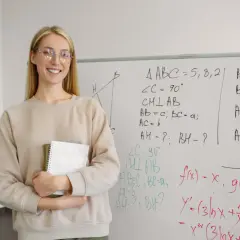 Uśmiechnięta nauczycielka z zeszytem przy tablicy z równaniami, w tym logarytmy rozszerzenie.